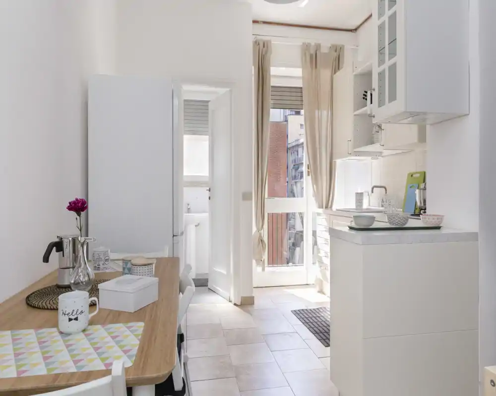 Compact, bright kitchen with a small breakfast table, countertop, and a door to a sunny balcony. Clean, modern styling with simple decor and natural light.
