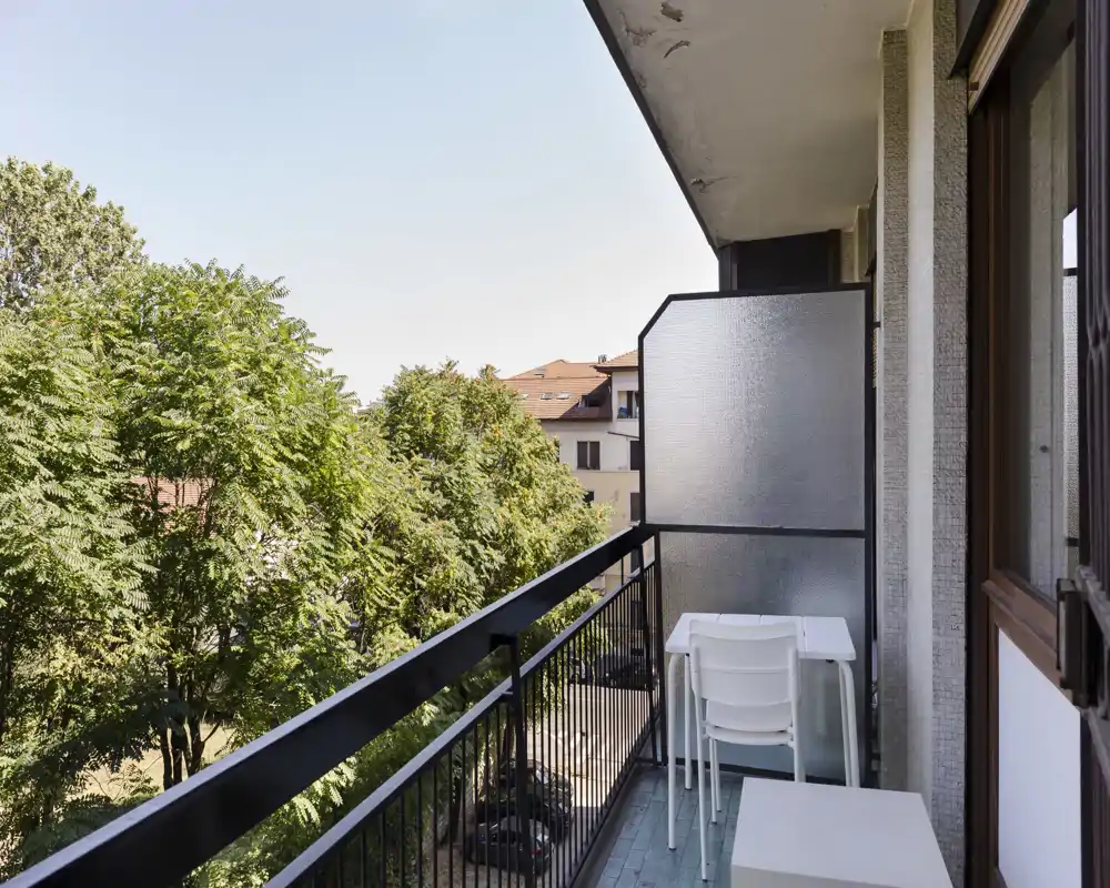 Sunny narrow balcony with a small table and stacked chairs, overlooking leafy trees and neighboring buildings — ideal for a morning coffee and light outdoor seating.