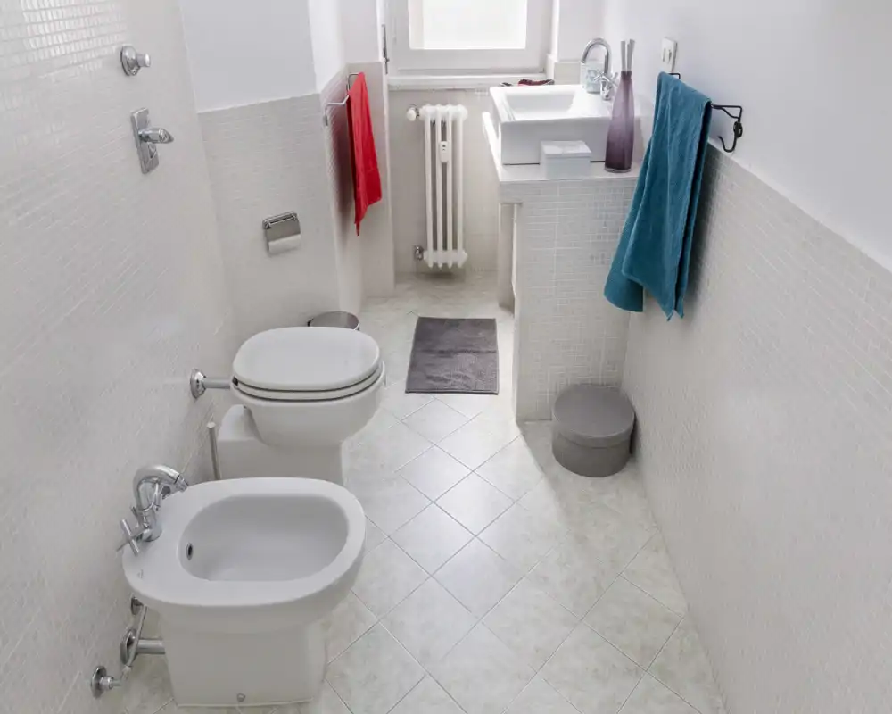 Bright, clean bathroom with a toilet, bidet and a sink on a tiled floor; neutral mosaic walls and colorful towels add a fresh touch.