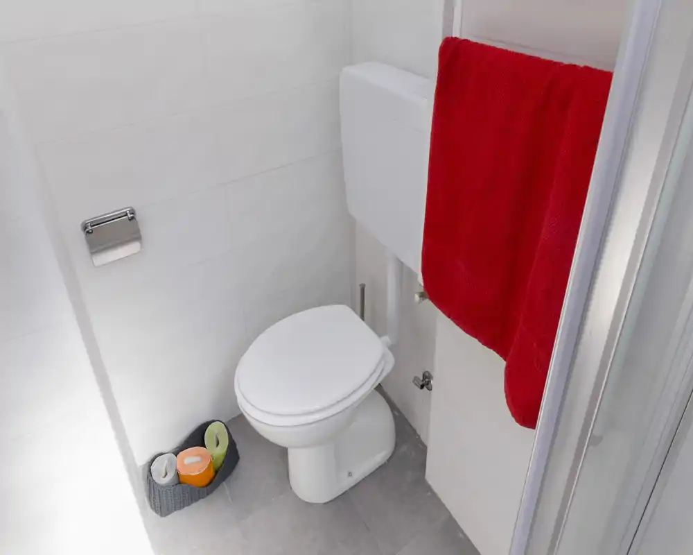 Compact, clean bathroom with a white toilet, tiled walls and floor, a red towel on a radiator and a small basket with toilet paper—bright and well maintained.