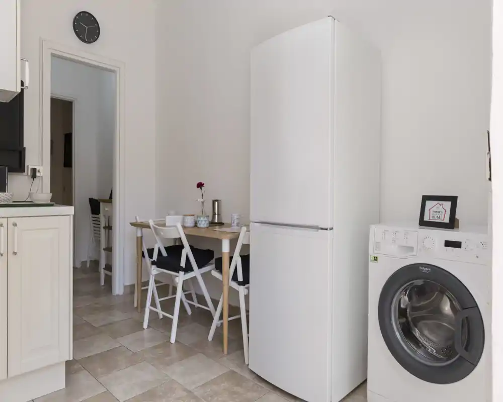 Bright, neatly styled kitchen corner with a full-height refrigerator, washing machine, and a small dining table with chairs — ideal for compact urban apartments.