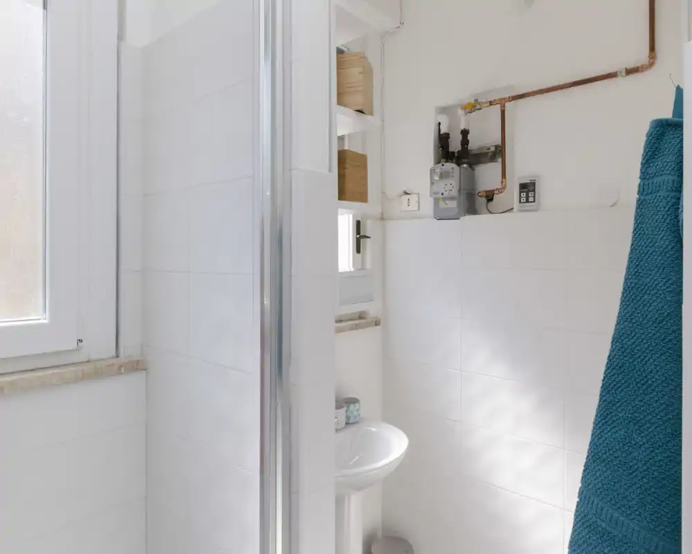 Bright, compact bathroom with white tiles, a pedestal sink and a shower screen visible; natural light enters through a frosted window.