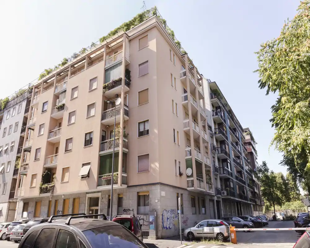 Exterior view of a multi-storey residential building with balconies, parked cars and street trees under clear daylight.