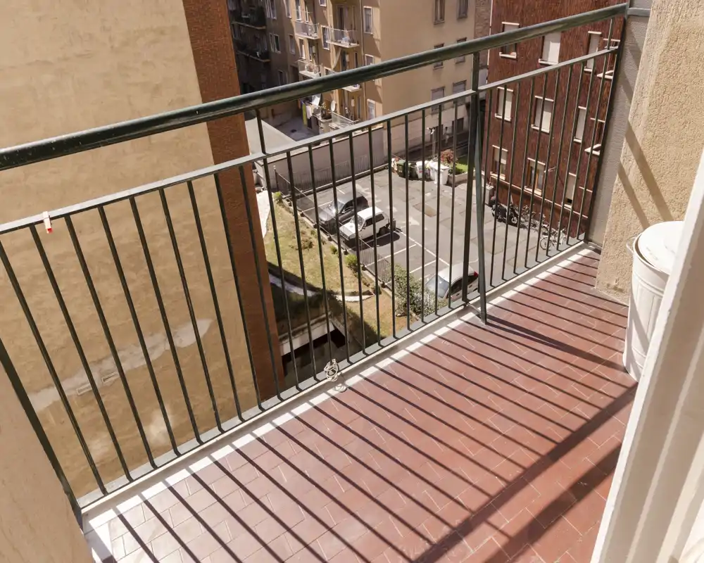 Sunny balcony with terracotta floor tiles and metal railing overlooking a residential courtyard; good natural light and space for a small table or plants.