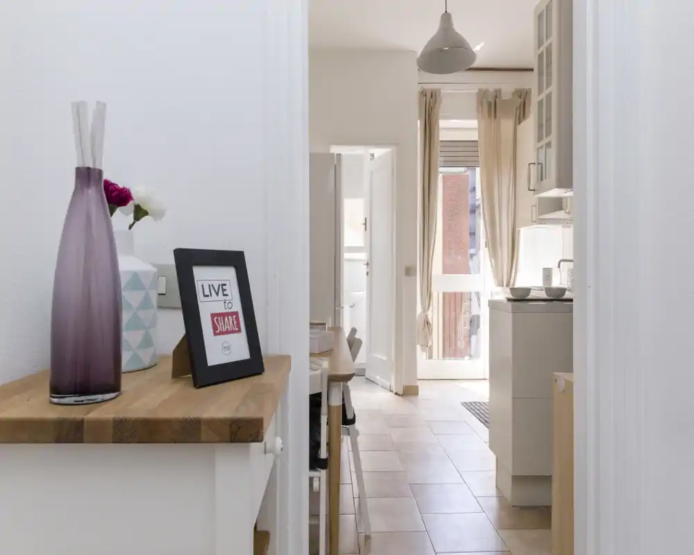 Bright, compact kitchen area with light tile flooring, a small island/counter and sunlight from a glass door. Decorative accents like a vase and framed sign give a welcoming touch.
