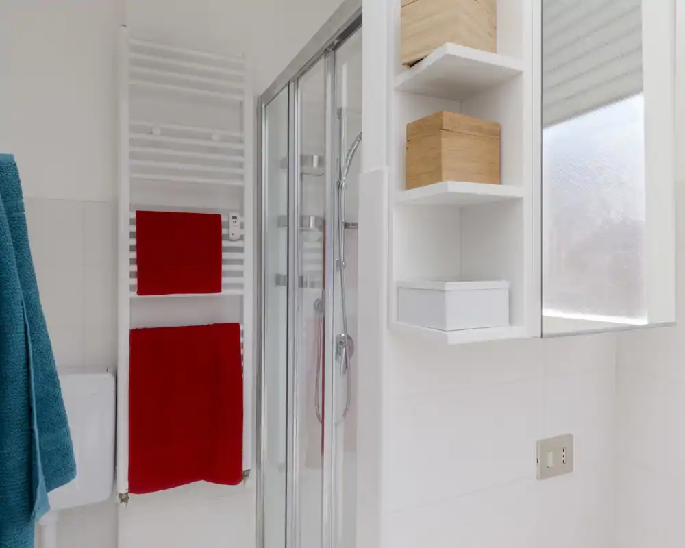 Clean modern bathroom with a glass-enclosed shower, white tiles, open shelving and colorful towels — bright and well maintained.