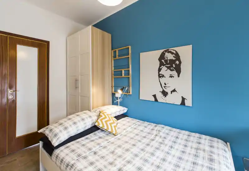 Dormitorio pequeño y luminoso con cama doble con ropa de cuadros, cojín amarillo decorativo, pared turquesa y armario — acogedor y moderno.