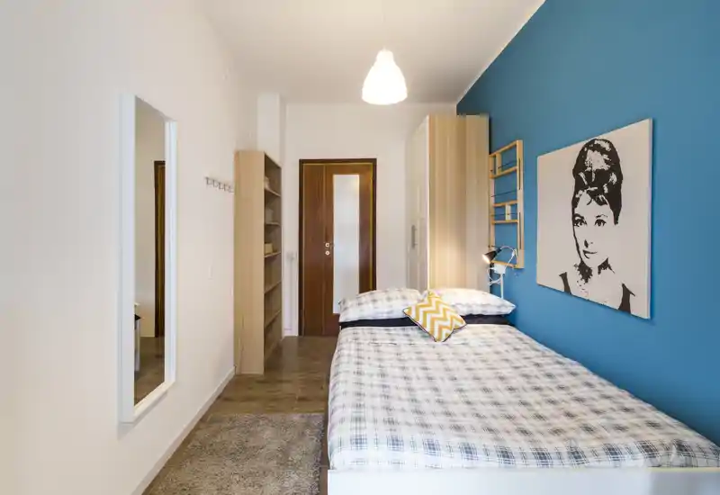 Dormitorio luminoso y bien decorado con colcha a cuadros, pared de acento turquesa, cuadro y armario; ideal para anuncios de alquiler que destaquen un espacio para dormir moderno y acogedor.