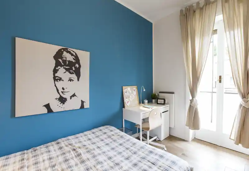 Dormitorio luminoso y moderno con pared turquesa, cama con ropa a cuadros, un pequeño escritorio blanco y una gran ventana que aporta mucha luz natural.