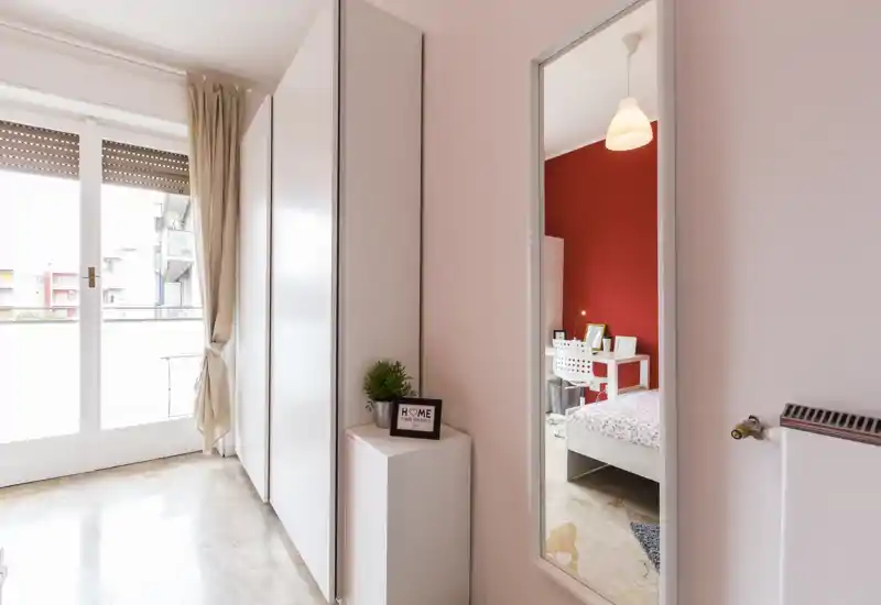 Dormitorio moderno y luminoso con puerta a balcón y espejo de cuerpo entero que refleja la cama y el escritorio; armario blanco y luz natural que crean una sensación ligera y minimalista.