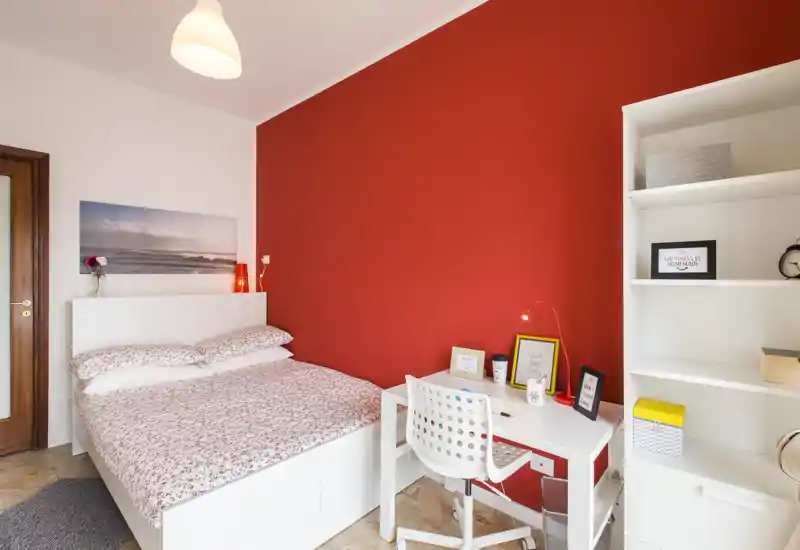 Dormitorio luminoso y ordenado con cama blanca, ropa de cama estampada y pared roja de acento. Un escritorio compacto y estanterías crean un espacio de trabajo práctico dentro de la habitación.