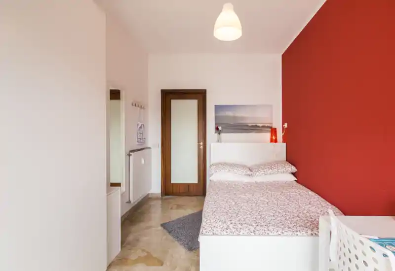 Dormitorio luminoso y compacto con cama doble junto a una pared roja intensa, mobiliario blanco y ropa de cama estampada. La habitación cuenta con lámpara, cuadro y una pequeña zona de escritorio — ideal para una estancia acogedora para uno o dos huéspedes.