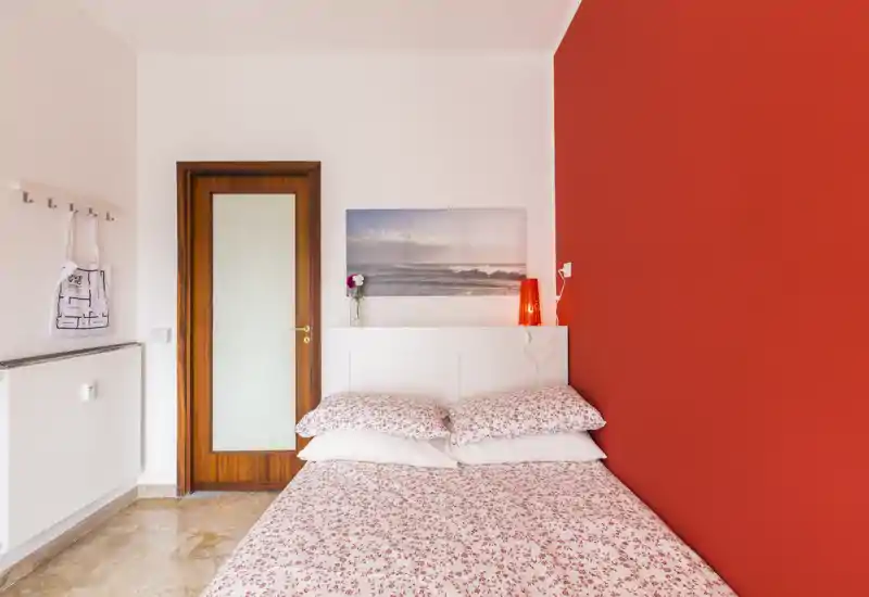Dormitorio luminoso y bien decorado con cama doble frente a una pared roja, ropa de cama floral, lámpara y cuadro costero — ambiente acogedor.