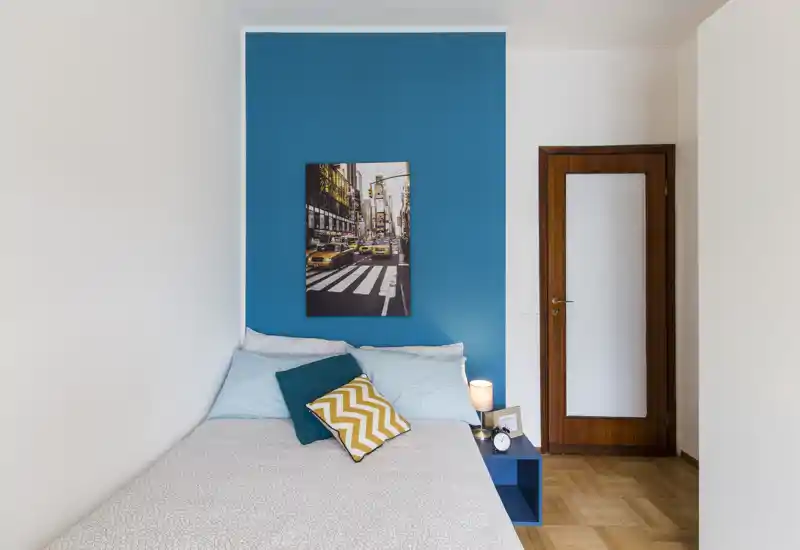Dormitorio luminoso y bien presentado con cama doble frente a una pared azul intensa, cojines decorativos y una mesita con lámpara — ideal para fotos de un anuncio.