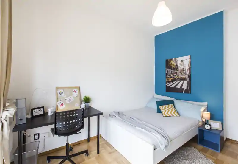 Dormitorio individual luminoso y moderno con pared azul de acento, cama cómoda, mesita con lámpara y un pequeño espacio de trabajo junto a la ventana — ideal para estudiante o profesional.