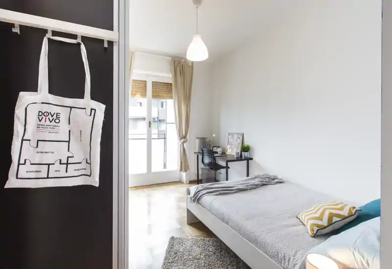 Dormitorio individual luminoso y bien decorado con cama baja, cojines estampados, pequeño escritorio junto a la puerta del balcón y alfombra suave — ideal para estudiante o profesional.