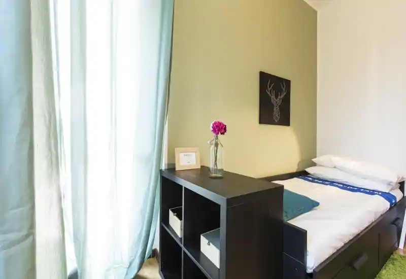 Dormitorio compacto y luminoso con cama individual, cortinas turquesa y estantería sencilla; luz natural agradable y decoración mínima, perfecto para estancias cortas.