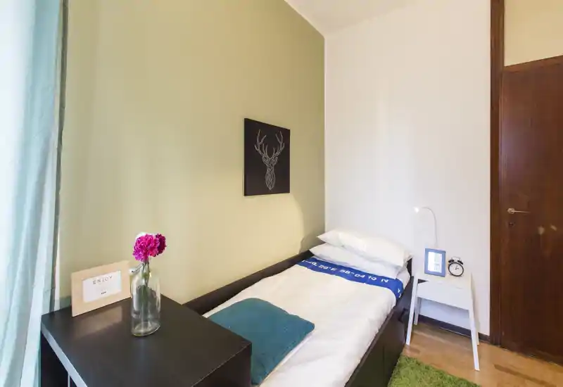 Habitación individual compacta y bien decorada con cama hecha, mesita de noche y luz natural desde la ventana. Decoración sencilla y toques cálidos que crean un ambiente acogedor y luminoso, ideal para estancias individuales.