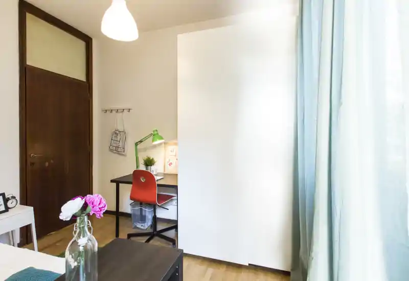 Rincón de dormitorio luminoso y bien cuidado con el borde de la cama visible, mesita de noche y un pequeño escritorio con silla roja, ideal para una sola persona. La luz natural entra por las cortinas claras y detalles como las flores y la lámpara de escritorio aportan calidez.