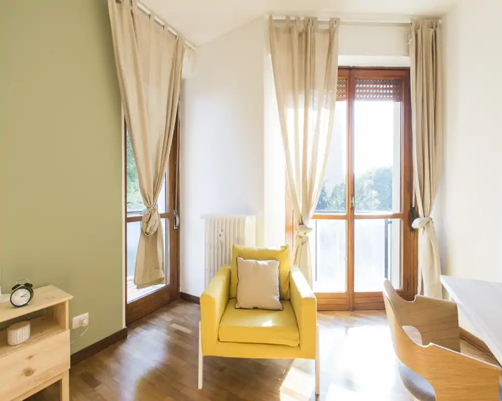 Zona de estar luminosa y acogedora con un sillón amarillo, cojín y grandes ventanas con cortinas ligeras que inundan la habitación de luz natural.
