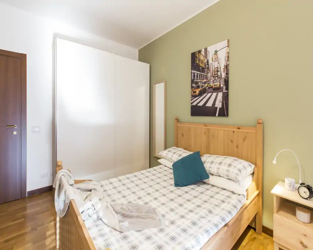 Dormitorio luminoso y ordenado con cama de madera, ropa de cama a cuadros y armario; la luz natural y el suelo de madera crean un ambiente acogedor.