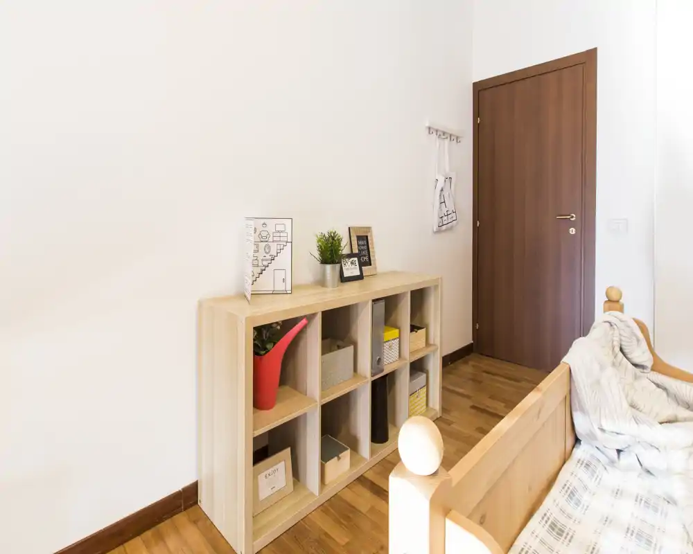 Rincón de dormitorio luminoso y ordenado con una cama individual de madera en primer plano y un mueble bajo con cajas decorativas y plantas contra una pared blanca.