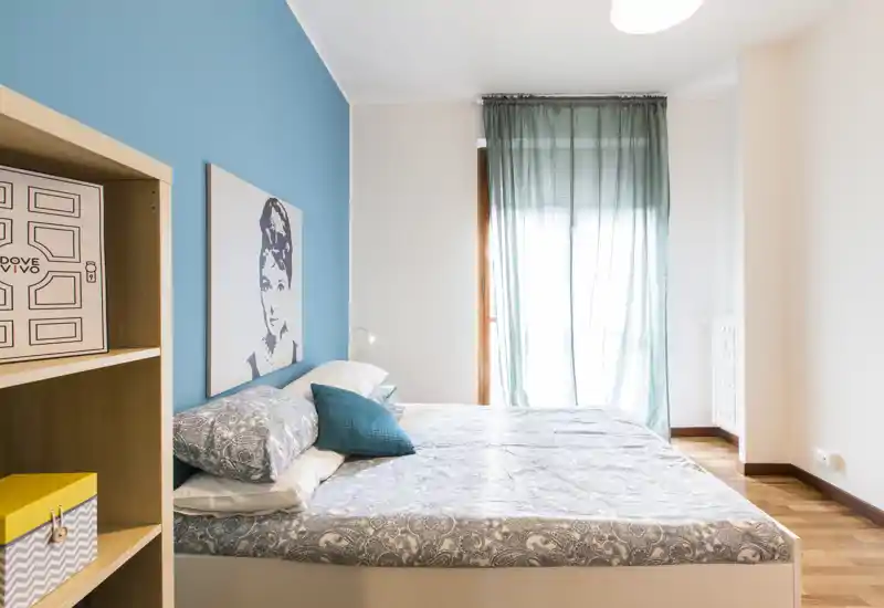 Bright, modern bedroom with a double bed dressed in patterned linens, teal accent pillows and a matching teal feature wall; sheer curtains let in soft natural light for a calm atmosphere.