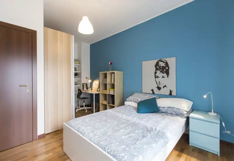 Bright, modern bedroom with a double bed against a blue accent wall, bedside table with lamp, shelving unit and a small work desk in the corner.