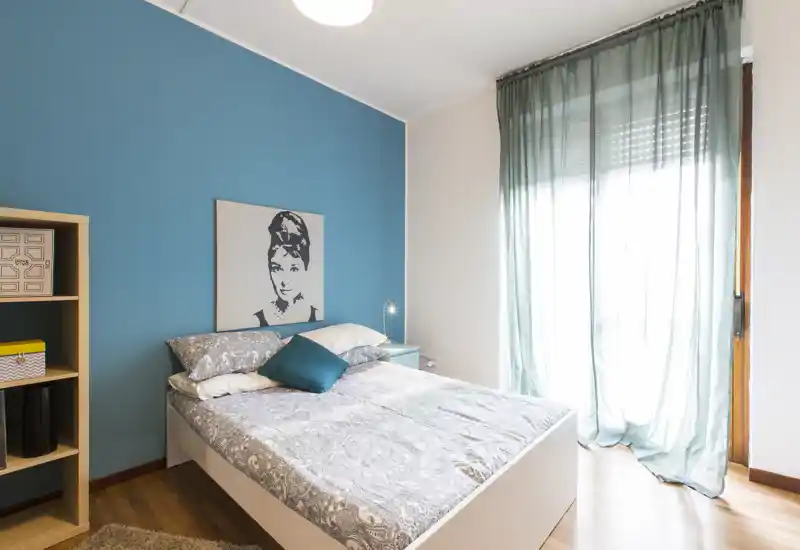 Bright, modern bedroom with a double bed, teal feature wall and large window letting in soft natural light — ideal for a relaxing stay.