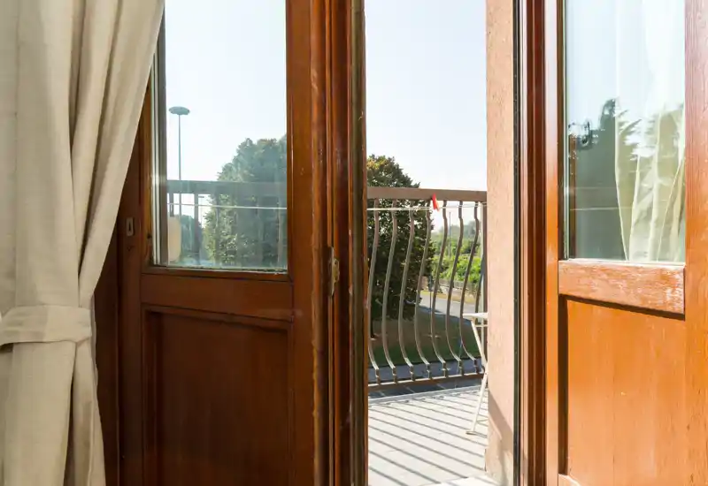 Petit balcon ensoleillé vu à travers des portes-fenêtres en bois ouvertes, avec une rambarde métallique et vue sur des arbres et une rue calme — idéal pour le café du matin.