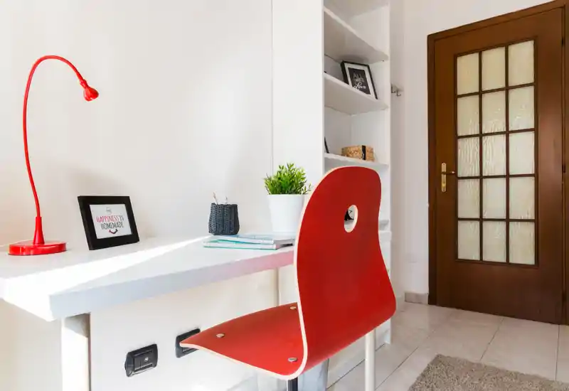 Coin bureau lumineux et minimaliste avec bureau blanc, chaise rouge et lampe, petite plante et étagères — adapté au travail ou à l'étude.