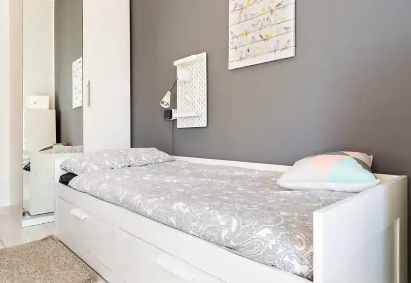 Camera singola luminosa e moderna con letto giorno bianco con cassettoni, biancheria dai motivi neutri e cuscino decorativo. Parete grigia d'accento, lampada da parete e guardaroba con anta a specchio che ampliano la luce e lo spazio percepito.