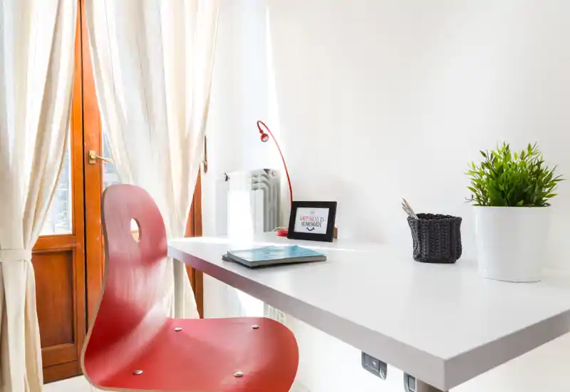 Coin bureau lumineux près d'une fenêtre avec chaise rouge, bureau blanc suspendu, petite lampe et plante — idéal pour travailler ou étudier dans un petit appartement.