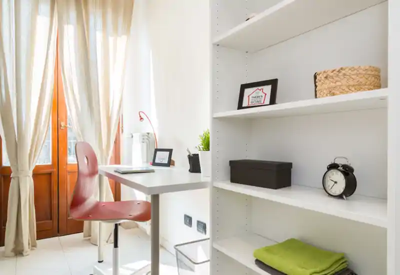 Espace de vie lumineux et polyvalent avec un petit bureau, une chaise rouge moderne et des étagères ouvertes — parfait comme coin bureau avec lumière naturelle venant des portes du balcon.