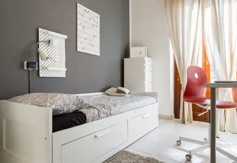 Camera singola luminosa e ordinata con un divano-letto bianco, tessuti morbidi e luce naturale dalla porta del balcone — ideale per un soggiorno accogliente.