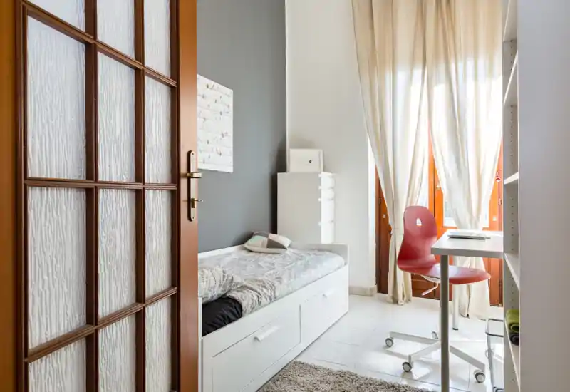 Camera singola luminosa e ben arredata con letto bianco con cassetti, piccolo scrittoio vicino alla finestra — ambiente accogliente e perfetto per un single o un ospite.