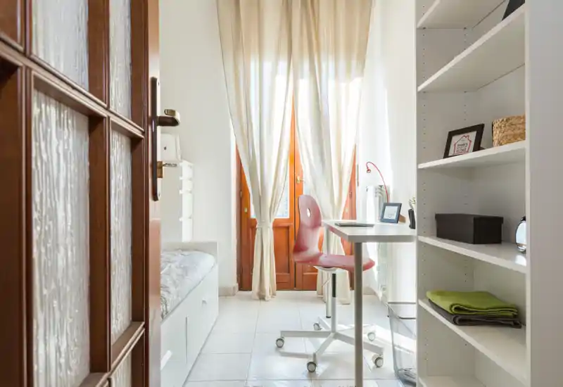 Camera singola luminosa e ben organizzata con scrivania vicino alla finestra. La luce naturale illumina la stanza, che dispone di scaffali e un letto compatto ideale per uno studente o un ospite.