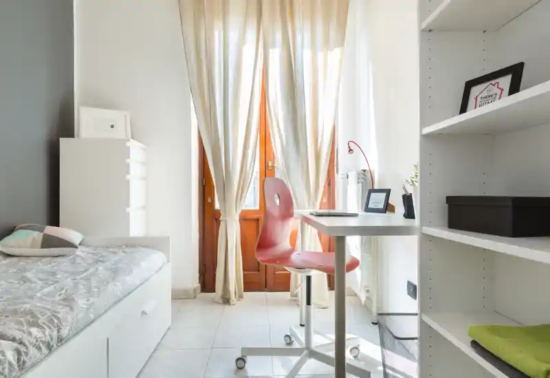 Bright, neatly styled bedroom with a single bed, study desk and shelving; natural light pours through tall curtains creating a calm workspace.