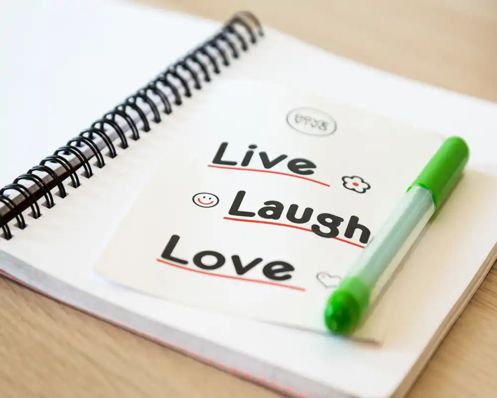 Primo piano di un taccuino a spirale con una penna verde e una card con la scritta "Live Laugh Love"; nessun ambiente o mobilio identificabile.