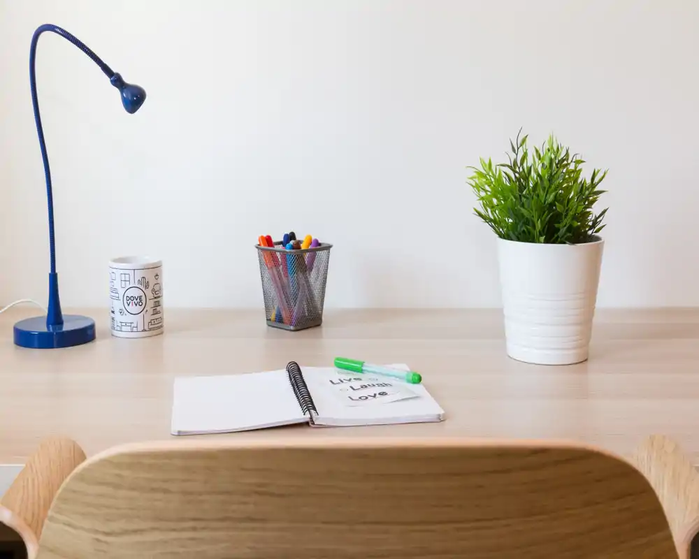 Scrivania luminosa e minimale con lampada, porta‑penne, taccuino e una pianta in vaso — ideale come angolo studio o home office.