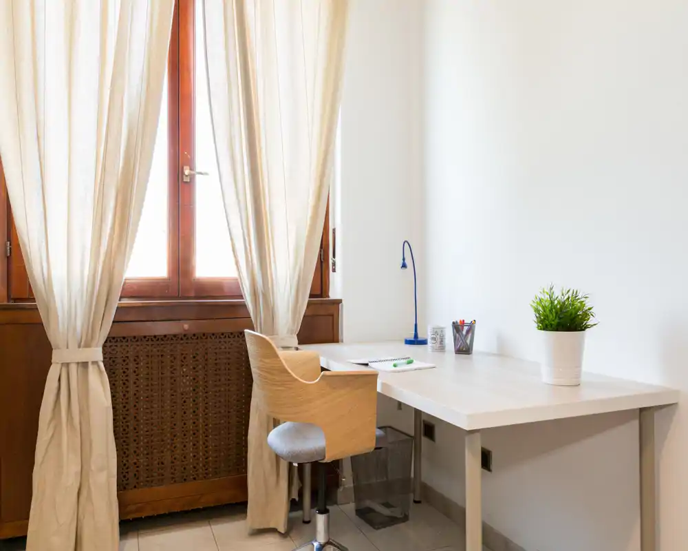 Angolo studio luminoso vicino alla finestra: scrivania bianca con sedia moderna in legno, lampada da tavolo, cancelleria e vaso con pianta. La luce naturale e le tende neutre creano un angolo tranquillo e ideale per il lavoro da remoto.