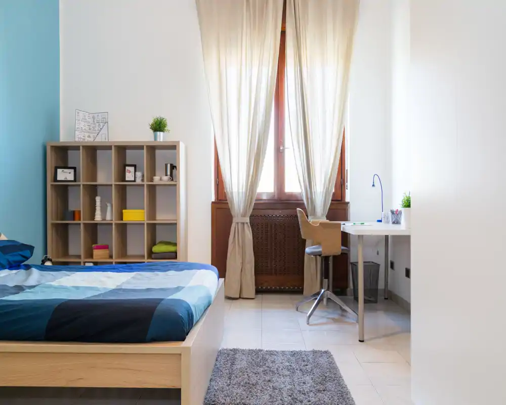 Camera da letto luminosa e ben arredata con letto in legno, libreria a cubi e scrivania vicino alla finestra che illumina l’ambiente con luce naturale.