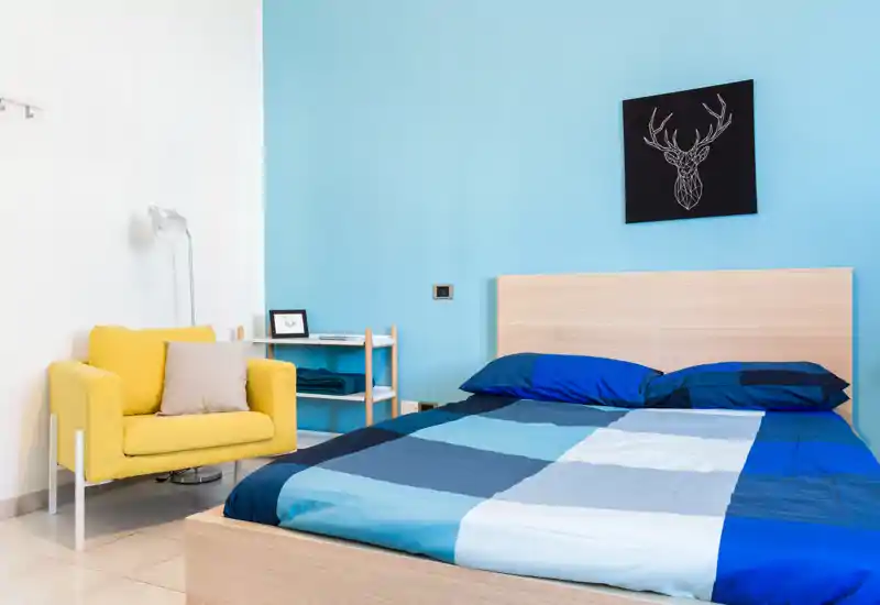 Camera da letto moderna e luminosa con letto matrimoniale con copertura blu a blocchi, testiera in legno chiaro e una poltrona gialla a contrasto, arredamento minimalista.