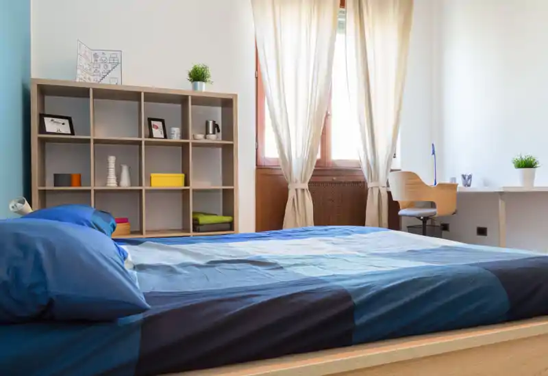 Camera da letto luminosa e ordinata con un grande letto in primo piano, luce naturale soffusa dalla finestra, e una libreria e scrivania minimal per riporre e lavorare.