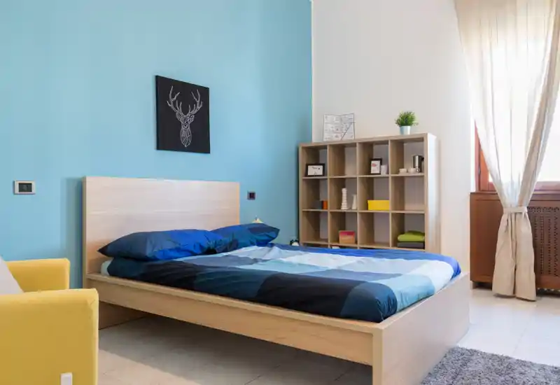 Camera da letto moderna e luminosa con letto matrimoniale in legno, biancheria blu e mensole decorative; luce naturale dalla finestra.