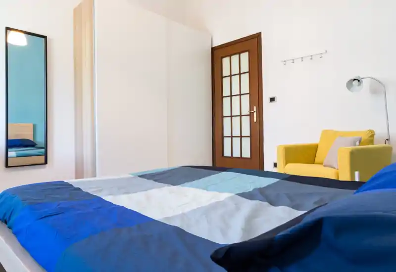 Bright, modern bedroom with a large bed covered in blue-gray linens, a white wardrobe, and a yellow armchair adding a pop of color. The space feels tidy and welcoming with natural light and simple, contemporary furnishings.