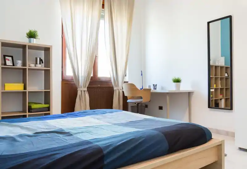 Camera da letto luminosa e ben arredata con un grande letto in legno in primo piano, una scrivania vicino alla finestra e scaffalature aperte per lo stoccaggio — ideale per un soggiorno confortevole.