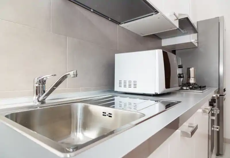 Cuisine moderne et compacte avec évier en acier inoxydable, plaque de cuisson intégrée et micro-ondes sur un plan de travail propre — lumineuse, pratique et idéale pour les locations courte durée.
