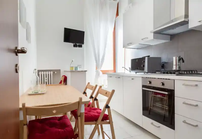 Kitchenette lumineuse et moderne avec placards blancs, four encastré et table en bois avec coussins rouges — idéale pour un séjour citadin compact.