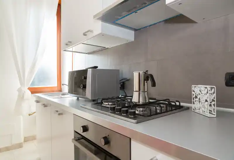 Cuisine moderne et compacte avec plaque de cuisson à gaz en acier inoxydable, four et une cafetière moka sur le plan de travail. La lumière naturelle d'une fenêtre rideau met en valeur les surfaces épurées et les appareils pratiques.